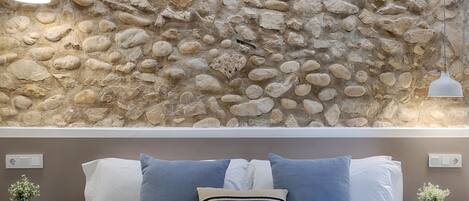 A bedroom with a stone wall, a bed with white and blue bedding, two bedside tables with plants, and pendant lights.