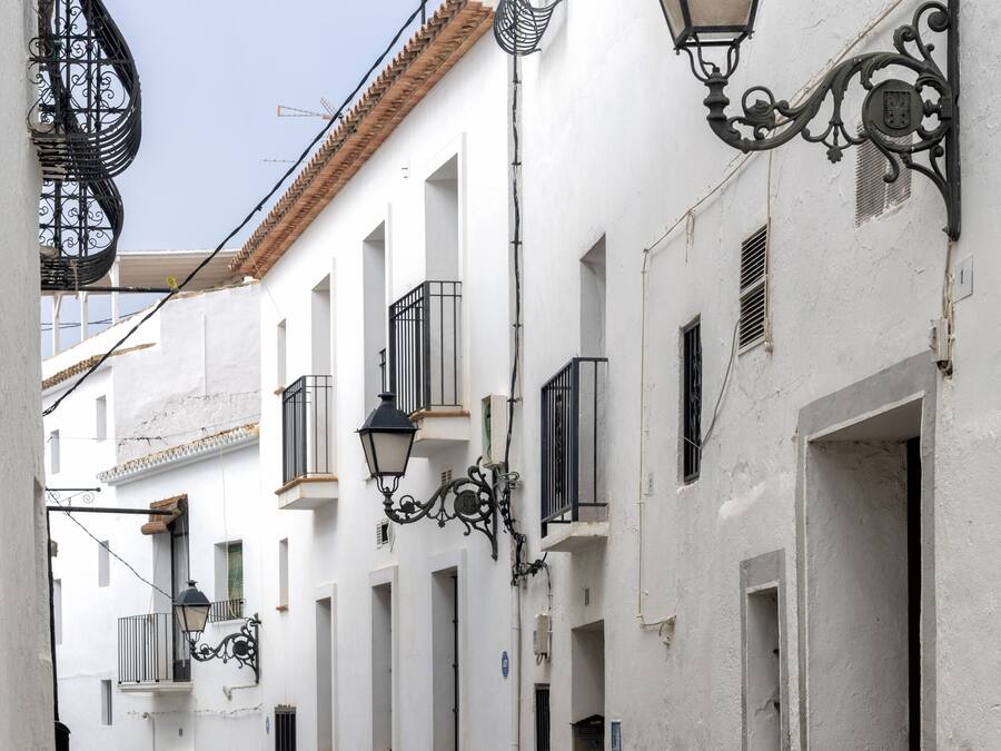 Una estrecha calle empedrada bordeada de edificios blancos con balcones de hierro forjado negro y farolas ornamentales.