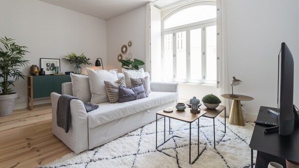 Un salon moderne avec un canapé blanc, une table basse et un téléviseur. On y trouve une fenêtre avec des rideaux, une plante et un meuble contenant des objets décoratifs.