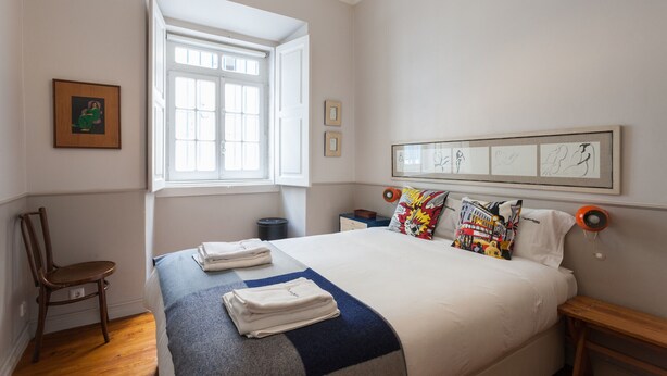 A neatly arranged bedroom with a large bed, a chair, a window, and a framed artwork on the wall. - FLH Janelas Verdes Charming Flat (Lisbon)