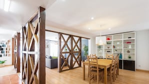 A modern interior with a wooden dining table and chairs, a white shelving unit with various items, and a cross-braced wooden partition.