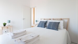 A neatly made bed with white linens and two pillows, a bedside table with a lamp, and a small plant.