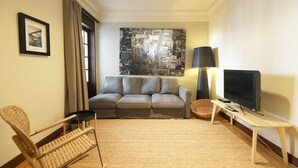 Una sala de estar moderna con un sofá gris, un sillón de mimbre, un televisor de pantalla plana sobre un mueble bajo, un cuadro de un paisaje urbano grande en la pared y una ventana con cortinas.