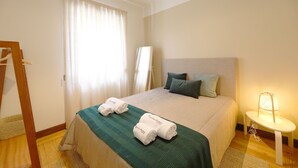 Habitación de hotel con cama, dos toallas, mesita de noche con lámpara y ventana con cortinas.