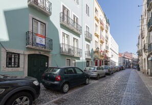 A narrow cobblestone street lined with multi-story buildings, parked cars, and balconies with metal railings.