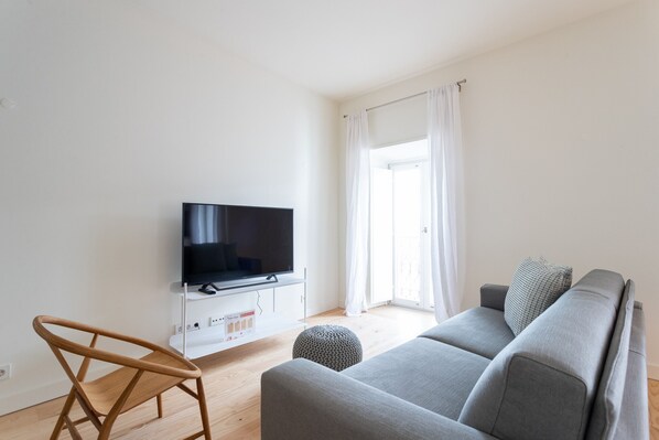 A modern living room with a flat-screen TV on a white stand, a grey sofa, a wooden chair, and a small ottoman.