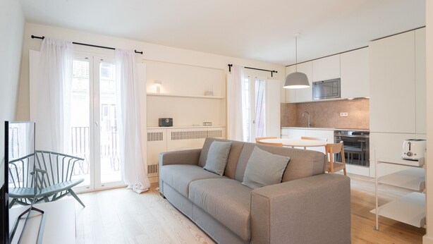 Ein modernes Wohnzimmer mit grauem Sofa, einem Esstisch und einer Küchenzeile mit weißen Schränken und Geräten.