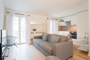 Un salon moderne avec un canapé gris, une table à manger et un coin cuisine doté de meubles et d’appareils électroménagers blancs.