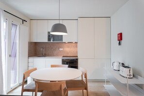 Une cuisine moderne avec des meubles blancs, une table de salle à manger ronde, des chaises en bois, un luminaire suspendu, un grille-pain et une machine à café.