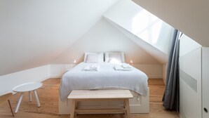 Ein kleines, modernes Schlafzimmer mit schrägem Dach, einem Bett mit weißer Bettwäsche, einem kleinen Tisch und einer Bank.