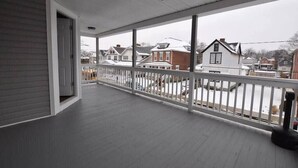 Une véranda couverte avec une balustrade blanche, un ventilateur de plafond et une vue sur les maisons voisines.