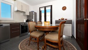 A compact kitchen with a dining area, featuring a round wooden table, four chairs, a refrigerator, a stove, and a window with blinds. - LOZA 2, 4+1 Sea View apt with Garden, 80m from sea (Slatine)