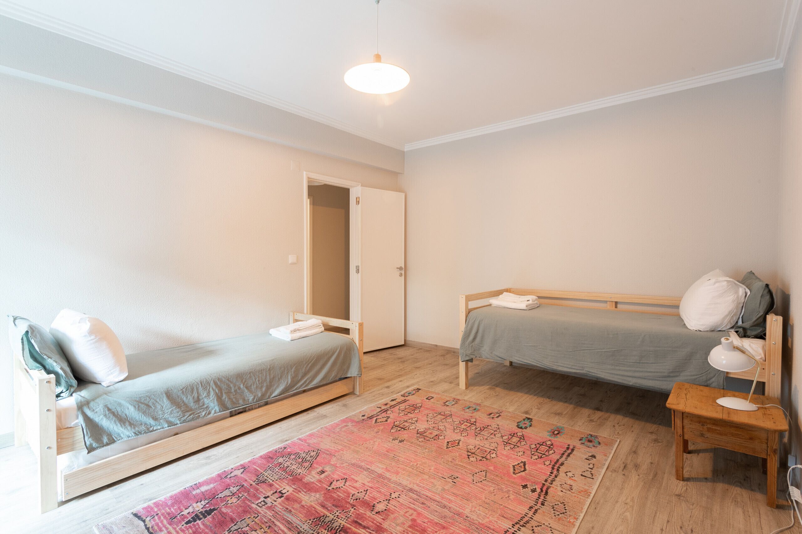 Habitación con dos camas individuales, suelo de madera, una puerta, una mesita con lámpara y una alfombra estampada.