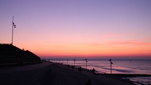En strand vid solnedgången med en klar himmel, en sandstrand och några få personer som promenerar.