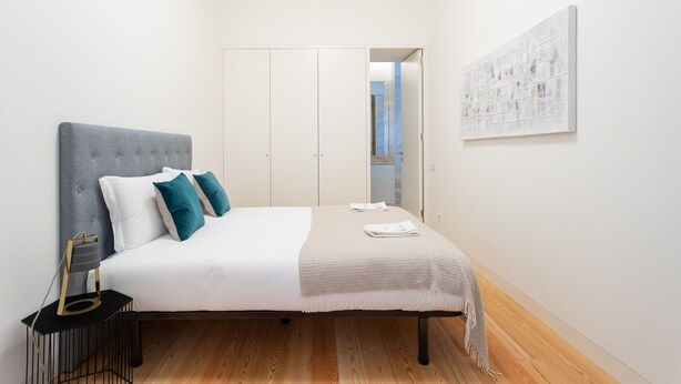 Une chambre moderne avec une tête de lit rembourrée de couleur grise, une literie blanche et des coussins turquoise. On y trouve une table de chevet avec une lampe, ainsi qu’une œuvre d’art encadrée accrochée au mur.