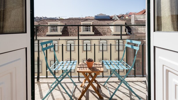 Ein Balkon mit zwei blauen Klappstühlen und einem Holztisch, mit Blick auf eine Stadtlandschaft mit terrakottafarbenen Dächern.