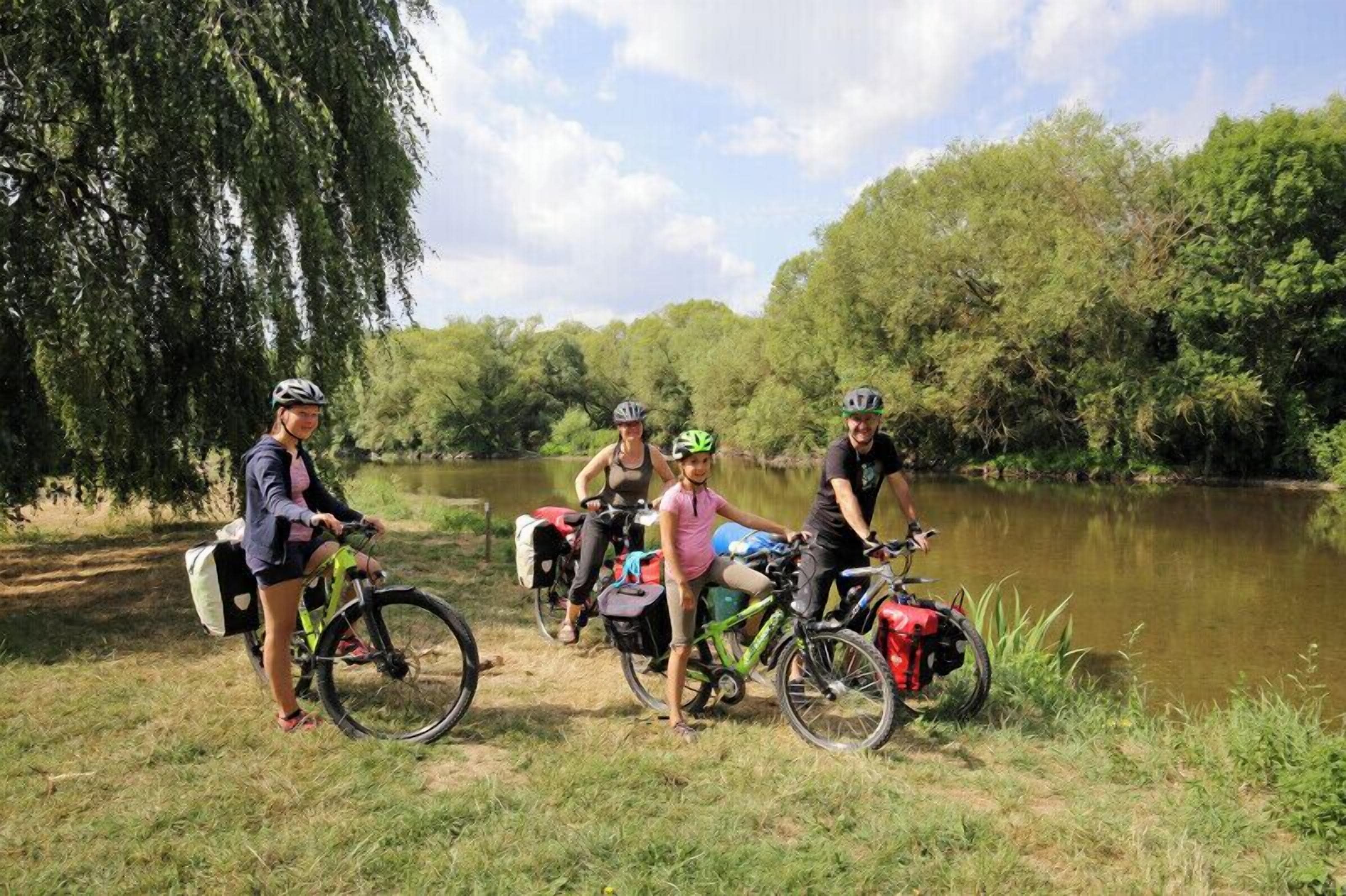 Four cyclists with bicycles near a body of water, surrounded by trees.