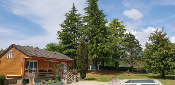 Una cabaña de madera con terraza, rodeada de árboles grandes y una zona cubierta de césped.