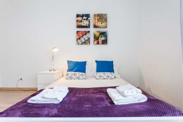 A neatly made bed with a purple blanket, two blue pillows, and two white pillows with a floral pattern. There are two folded towels on the bed. A bedside lamp is on the nightstand beside the bed. Four artwork pieces are hung on the wall above the bed.