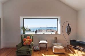 A cozy living room with a large window overlooking a cityscape, a green armchair, a small table, and a television.