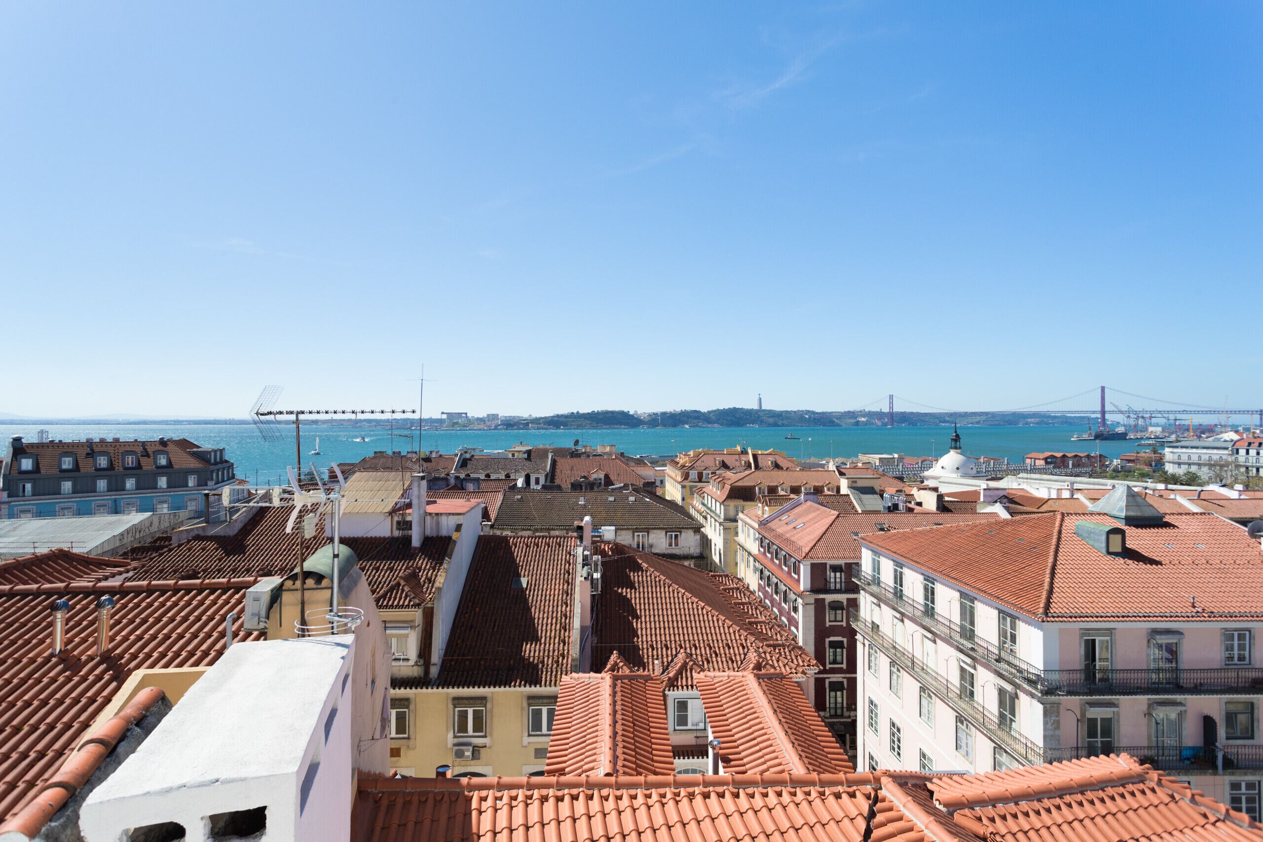 A cityscape with terracotta-roofed buildings, a prominent bridge in the background, and a clear blue sky.
