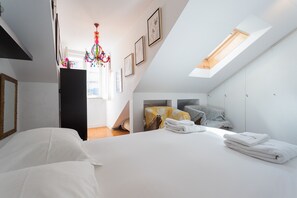 Une chambre moderne avec une lucarne, un lit recouvert de draps blancs, un lustre coloré et des tableaux encadrés accrochés au mur.