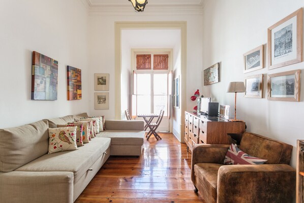 Ein Wohnzimmer mit einer Couch, Sesseln, einem Couchtisch und einem TV-Schrank. An den Wänden hängen Bilder in Rahmen und ein Fenster mit Rollläden.