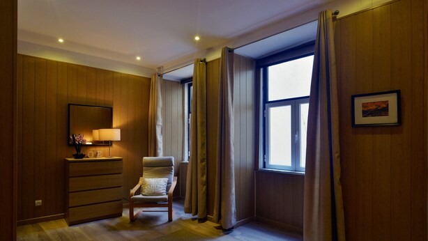 Ein Hotelzimmer mit Holzwandverkleidung, einem Bett, einem Sessel, einer Kommode, einem Fenster mit Vorhängen und einem gerahmten Bild an der Wand.