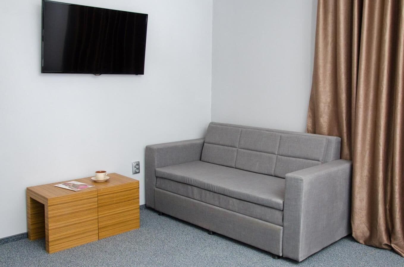 A grey sofa, a wooden side table with a cup, and a TV on the wall.
