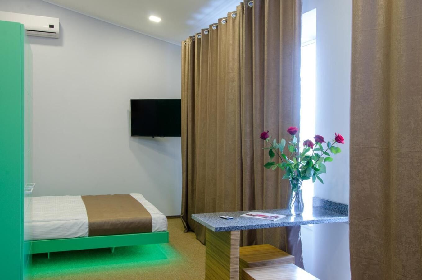 A hotel room with a bed, a TV mounted on the wall, a small table with a vase of flowers, and a window with curtains.