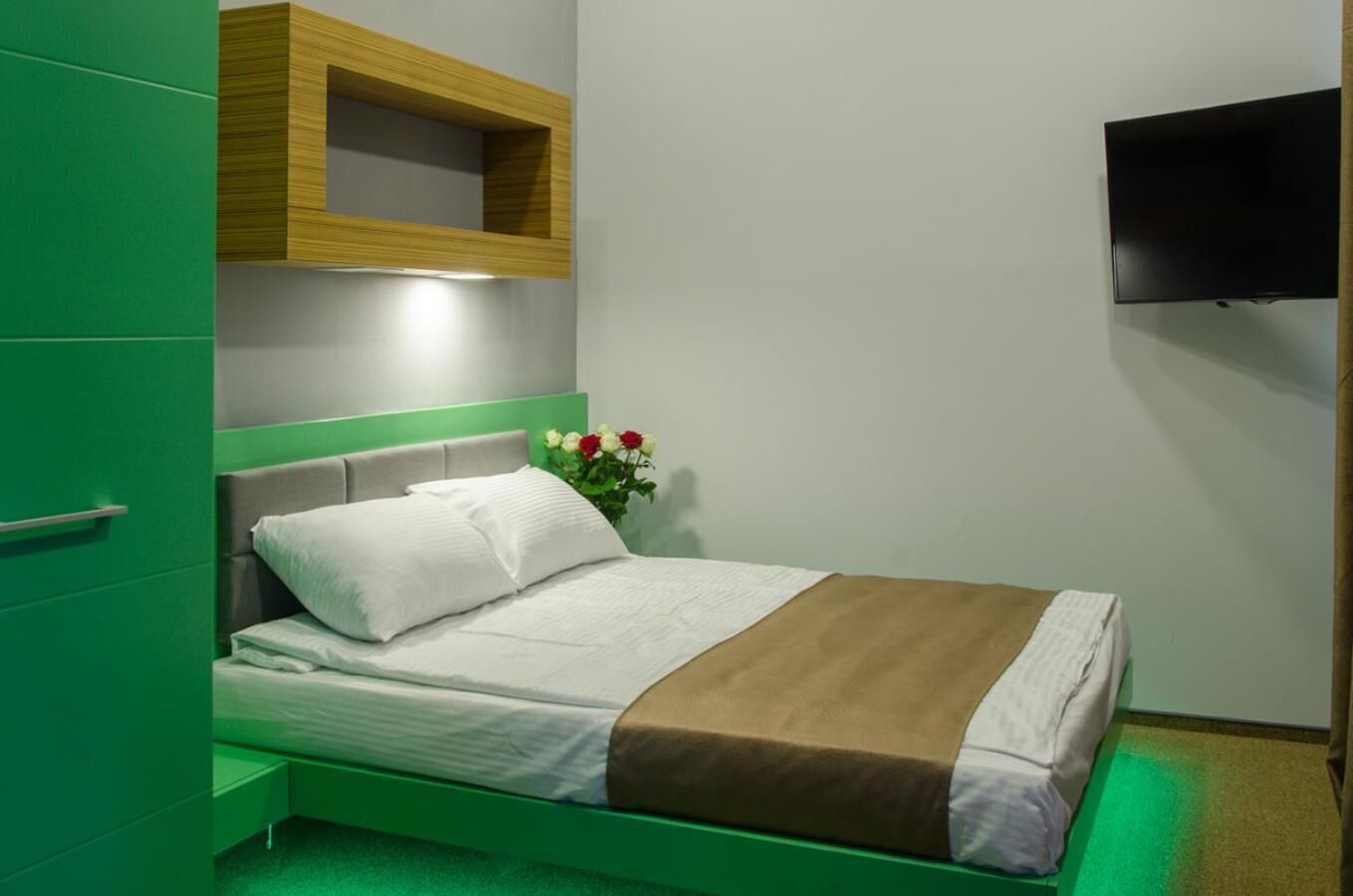 A modern bedroom with a green bed, a wooden shelf above the bed, a flat-screen TV on the wall, and a vase with flowers on the nightstand.