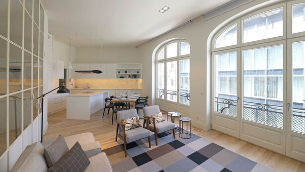 A modern living room with a sofa, armchairs, and a coffee table. There is a kitchen area with a dining table and chairs. Large windows provide a view of the outside. - FLH Sá da Bandeira Luxury Apartment (Oporto)