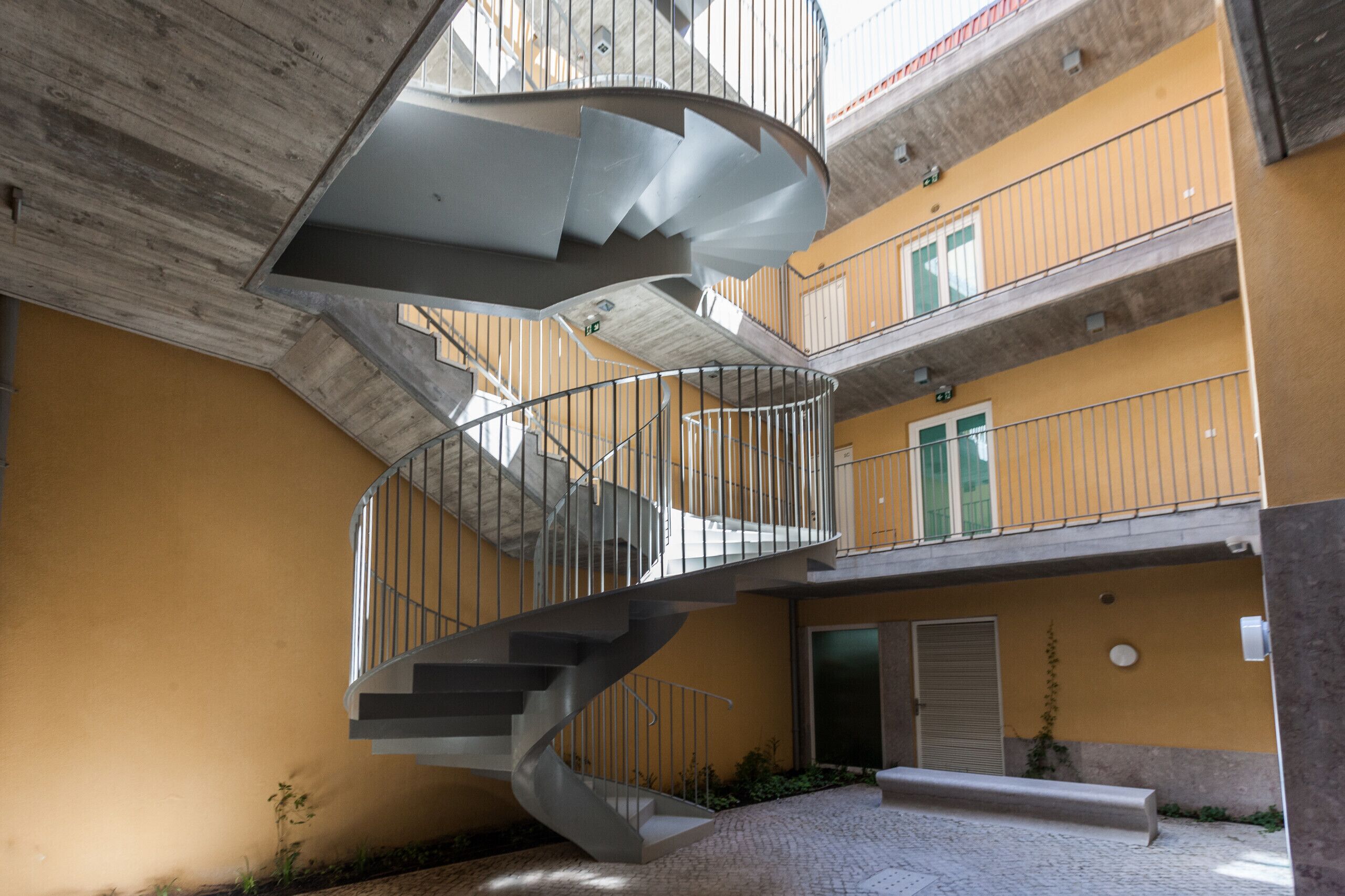 Una scala a chiocciola su più livelli, con ringhiere in metallo, collega i diversi piani di un edificio con pareti gialle e soffitto in cemento.