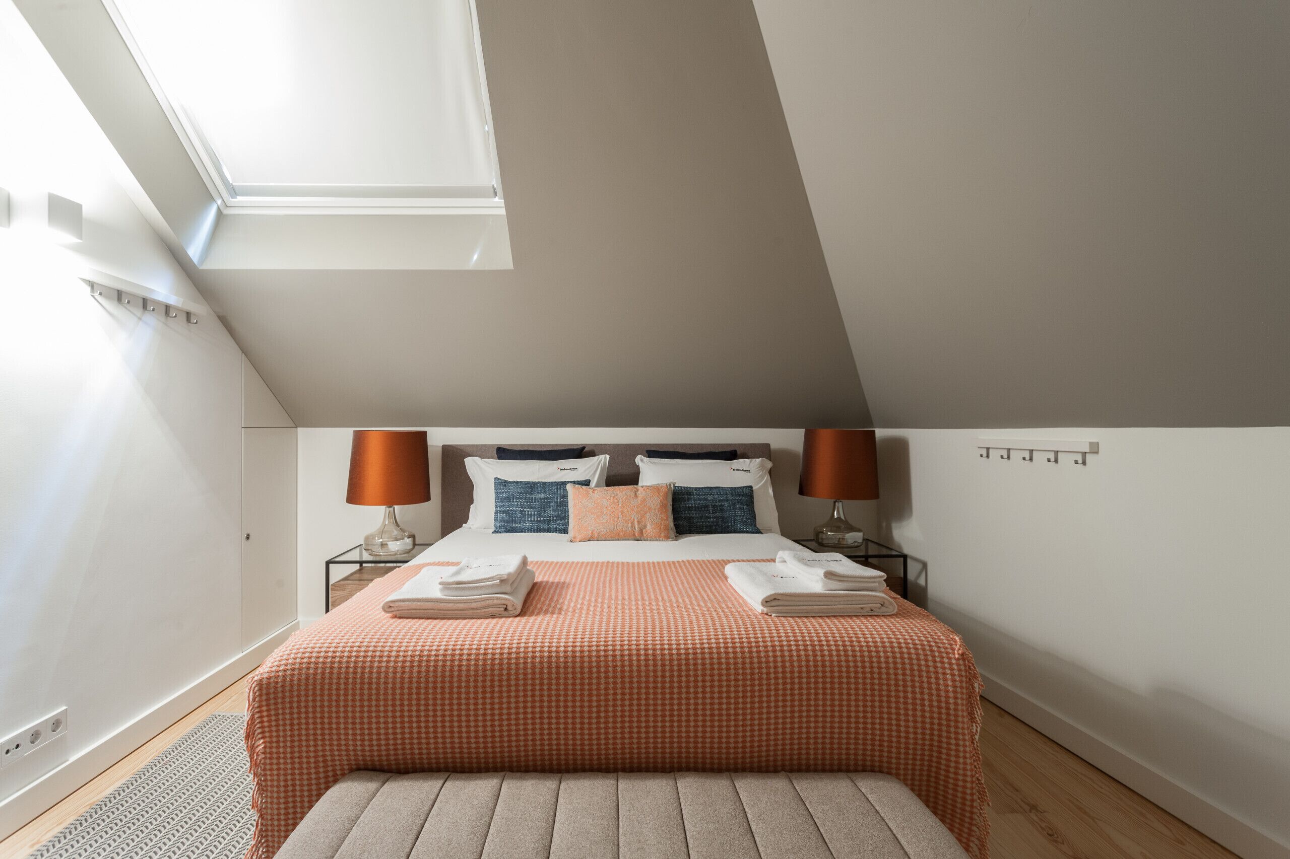 Una camera da letto moderna con un lucernario, un letto rifatto con copriletto a quadri, due abat-jour e un piccolo divanetto ai piedi del letto.