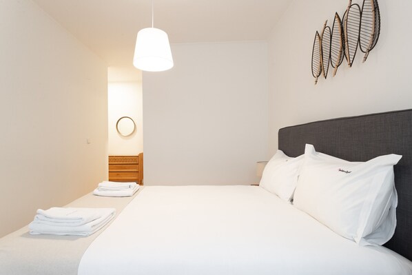 A modern hotel room with a large bed, white linens, a grey headboard, a hanging light fixture, and a wall-mounted decorative item.