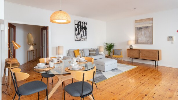 Ein modernes Wohnzimmer mit Essbereich, ausgestattet mit einem runden Glastisch, Holzstühlen, einer Couch und einem großen abstrakten Gemälde an der Wand.