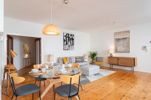 A modern living room with a dining area, featuring a round glass table, wooden chairs, a sofa, and a large abstract painting on the wall.