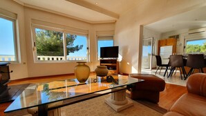 A spacious living room with a glass coffee table, a sofa, and a TV. There are large windows with a view of trees and a balcony. - Villa Blue Adriatic (Omis)