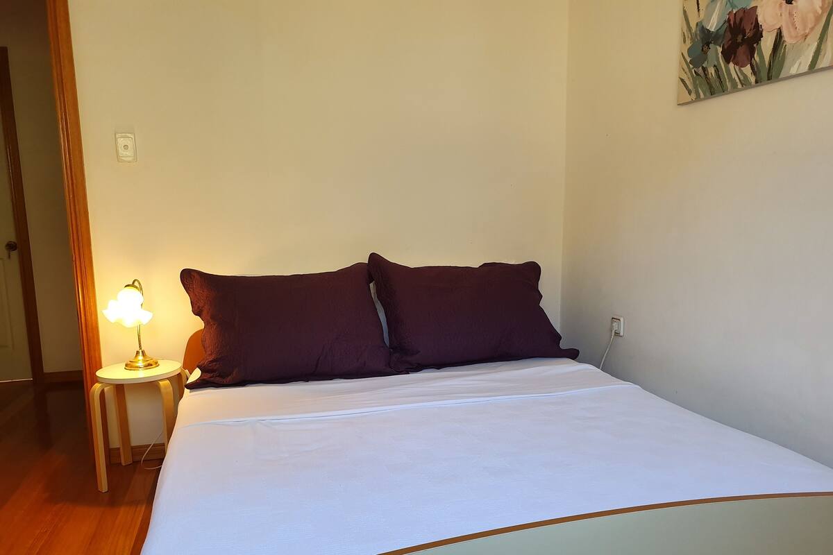 Una cama bien hecha con ropa blanca y dos almohadas oscuras, una lámpara de noche con pantalla en forma de nube y un enchufe en la pared.