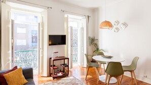 En stue med sofa, fjernsyn, spisebord med stole og en balkon med gelænder.