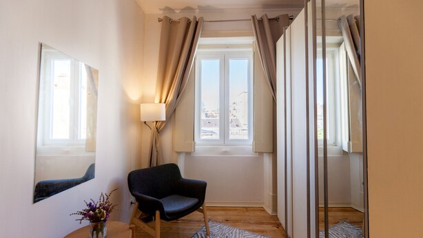 Et værelse med en stol, et lille bord med en vase, en lampe og et vindue med gardiner.
