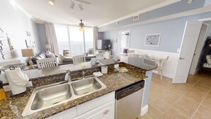 A modern kitchen with granite countertops, stainless steel sinks, and a dishwasher. There is a dining area with a table and chairs, and a balcony with a view. - Master on Gulf -Free round of Golf, Beach Chairs, & Activities Every Day! (Panama City Beach)