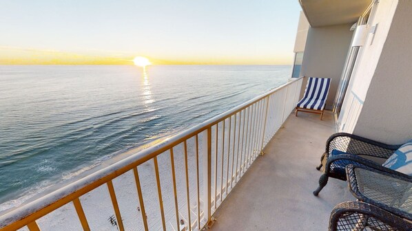 A balcony with a view of the sea at sunset, a striped chair, and a wicker chair. - Master on Gulf -Free round of Golf, Beach Chairs, & Activities Every Day! (Panama City Beach)