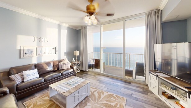 A modern living room with a sofa, coffee table, and TV. It features a balcony with a view of the ocean. - Master on Gulf -Free round of Golf, Beach Chairs, & Activities Every Day! (Panama City Beach)