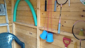 Une pièce en bois avec des raquettes de badminton, un tuyau d’arrosage vert et une chaise en plastique bleue.