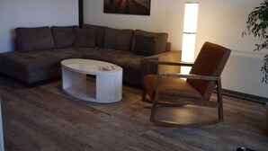 Un salon comprenant un canapé d’angle, une table basse en bois, un fauteuil en bois et un tableau représentant un phare accroché au mur.