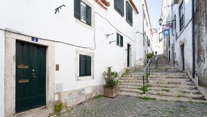 Una estrecha calle adoquinada bordeada por edificios blancos con contraventanas verdes, una puerta de madera con el número 3 y una pequeña planta en maceta.
