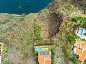An aerial view of a coastal property with a swimming pool, a house with a tiled roof, and a rocky cliff.