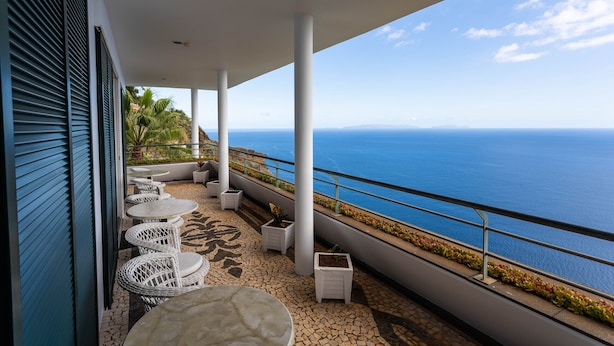 Een balkon met witte wicker stoelen en tafels, een betegelde vloer en uitzicht op de oceaan.