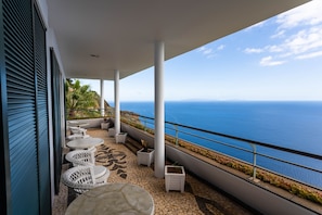 A balcony with white wicker chairs and tables, a tiled floor, and a view of the ocean.